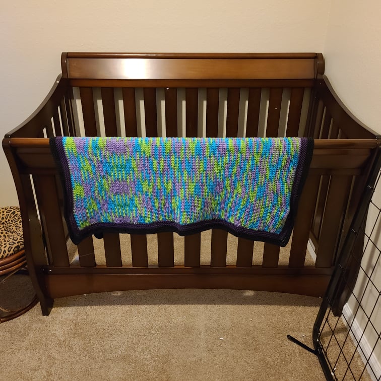 Newly assembled crib frame with a blue, purple, and green crocheted blanket that has a double border of dark purple and black.